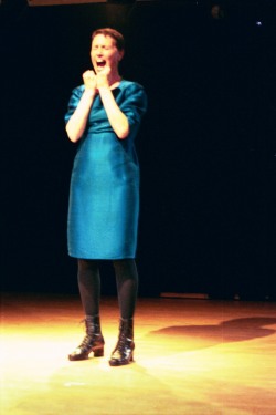 Adina with her hands in fists near her mouth , which is open - as if she were yelling.  Her eyes are closed.  She is wearing a blue dress, black boots and standing on a yellow floor.